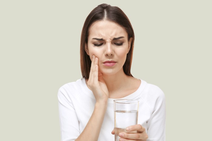 A young woman experiencing tooth sensitivity after using teeth whitening products, holding her jaw with one hand and a glass of water in the other.