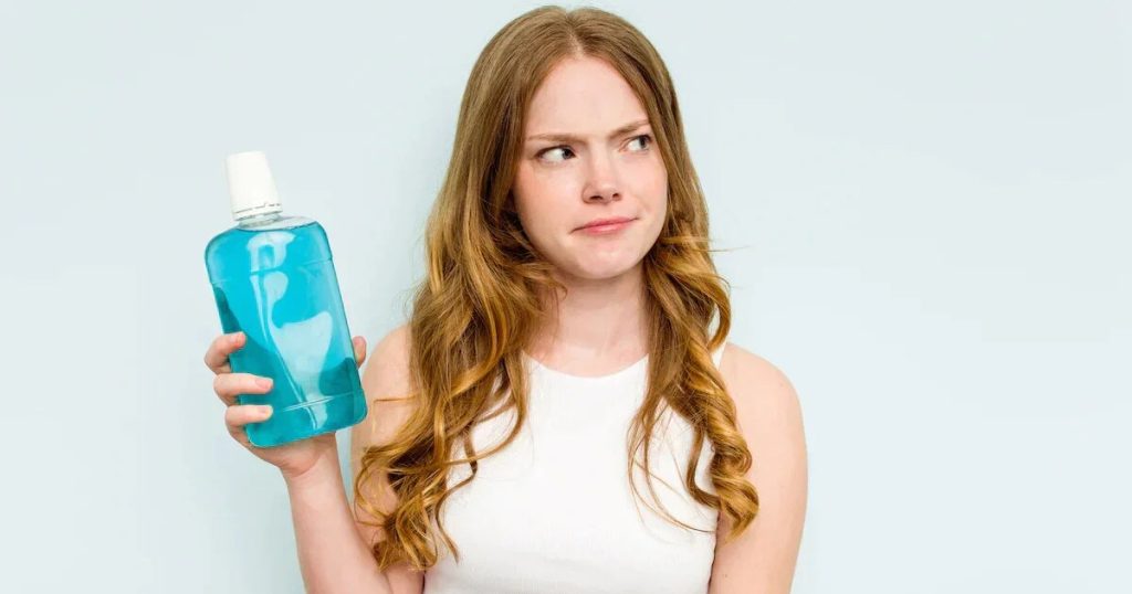 A young woman holding a mouthwash bottle in her hand with a hesitant look on her face.