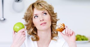 Young woman thoughtfully holding two different foods in her hands, deciding what to eat after root canal.
