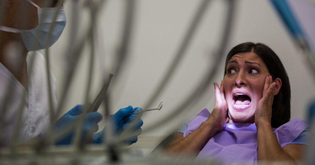 A frightened woman sitting in a dental chair screaming while a dentist stands beside her, illustrating severe dental phobia.
