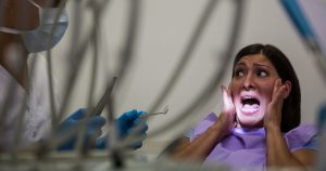 A frightened woman sitting in a dental chair screaming while a dentist stands beside her, illustrating severe dental phobia.