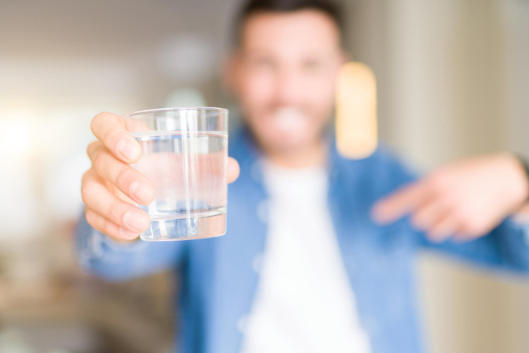 Man pointing at a clear glass of water in focus, blurred background, concept of hydration and dry mouth relief