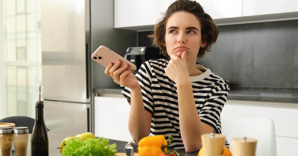 Young woman hesitating in the kitchen, surrounded by food, unsure when she can eat solid food after tooth extraction.
