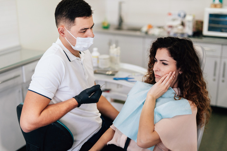 Upset young woman in dental chair after tooth damage during treatment, dentist standing nearby in clinic
