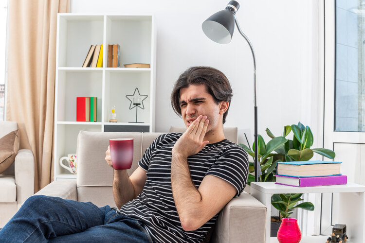 A young man sitting at home, experiencing tooth pain after a dental cleaning procedure.